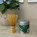 Bamboo whisk and green tea canister on a wooden surface with a plant in the background